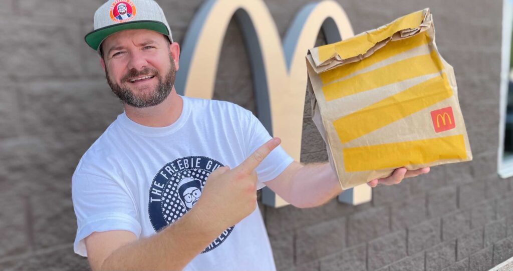 The Freebie Guy holding a McDonald's sack and pointing at it standing in front of a McDonald's store