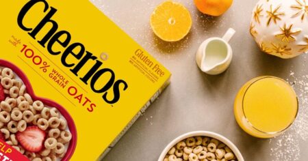 Box of Cheerios laying on a table with a bowl of Cheerios next to it