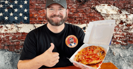 Kendall holding a Taco Bell pizza at table with American Flag brick wall